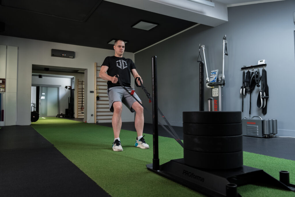 Trener personalny Dawid Dzikiewicz podczas treningu z sańmi obciążonymi talerzami w profesjonalnym studio.